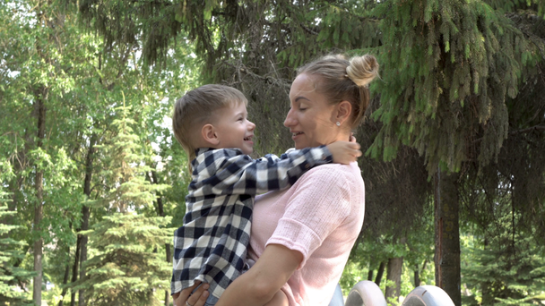 Szczęśliwa młoda matka trzyma dziarski, śmiejąc się dziecko w ramionach. Przytulić i przytulać głowę, aby udać się na tle zielonych drzew w parku miejskim. Trochę żartuje chłopiec i przeraża kamerzystę. 4K - Materiał filmowy, wideo