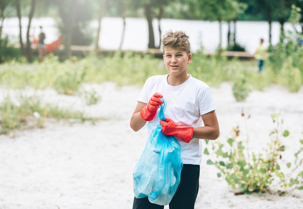 Boy in white t shirt in gloves collects garbage and plastic bottles into blue package on the beach. Young volunteer. Environmental protection, save environment concept.  - Foto, Imagen