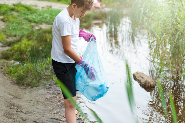 Το αγόρι με το λευκό μπλουζάκι στα γάντια συλλέγει σκουπίδια και πλαστικά μπουκάλια σε μπλε πακέτο στην παραλία. Νέος εθελοντής. Προστασία του περιβάλλοντος, αποθήκευση της έννοιας του περιβάλλοντος.  - Φωτογραφία, εικόνα
