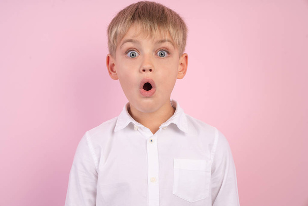 Surprise, excitement and fascination concept. Funny bug eyed little boy opening his mouth widely, shocked with astonishing unexpected news, having amazed look, showing full disbelief. isolated on pink - Photo, Image