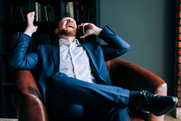 Úspěšný podnikatel v obleku sedí v křesle módní kanceláře a směje se nahlas. - Fotografie, Obrázek