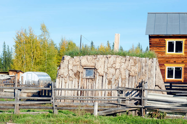 Комната для животноводства, сделанная из коровьего навоза и древесины, заросшая травой стоит на современном участке с домом на севере Якутии
. - Фото, изображение
