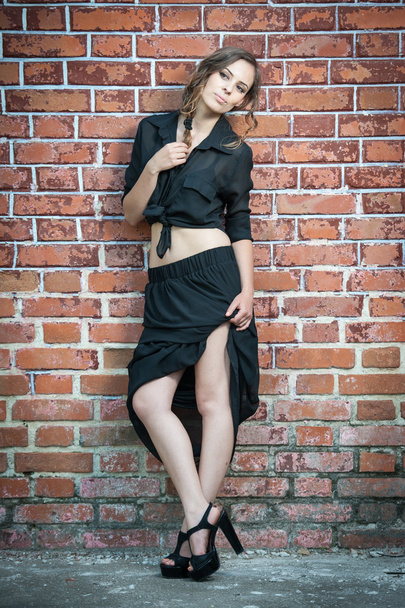 Charming young brunette woman in black dress and high heels near the brick wall.Sexy gorgeous young woman near old wall.Full length portrait of a cute woman with long hair near a brick wall - Zdjęcie, obraz
