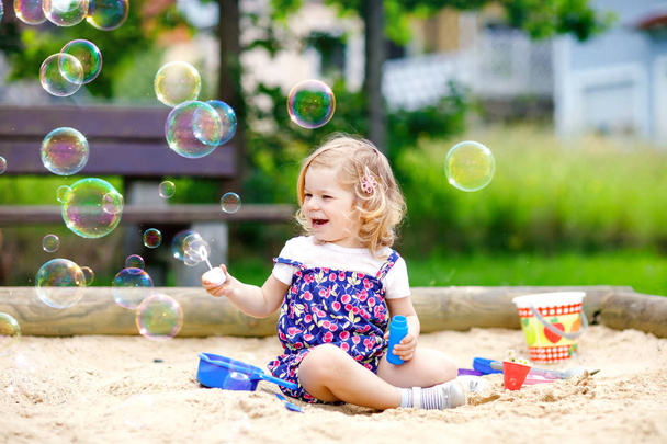 Beautiful little blonde toddler girl having fun with blowing soap bubble blower. Cute adorable baby child playing on playground on sunny summer day. Happy active funny healthy kid - Photo, Image