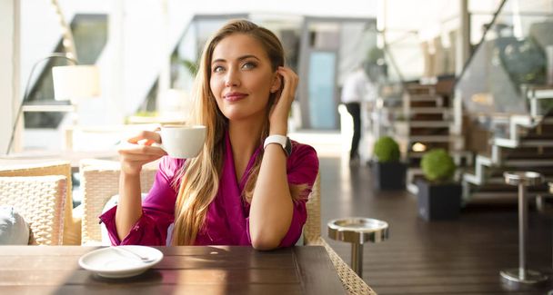 Attractive young model in a cafe drinking tea in the summer morning. Sun is shining, warm day. Wearing pink silk dress. Modern makeup and stylish hair. Touching ear. Watch on the wrist. - 写真・画像