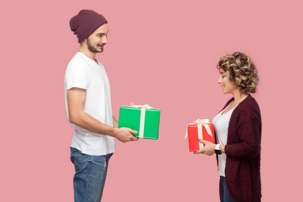 Side view of surprised couple of friends in casual style giving present boxes to each other while celebrate anniversary on pink background - Fotoğraf, Görsel