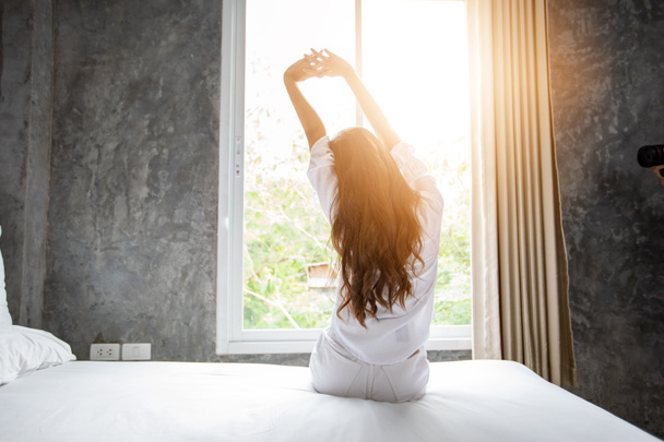 ベッドで目を覚ますアジアの女性は、新鮮な空気で朝に開いたカーテンで完全に休んだ. - 写真・画像