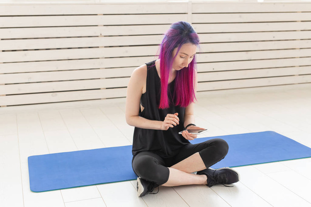 Abbastanza giovane ragazza mista con i capelli viola sta contando calorie bruciate utilizzando un'app sul suo smartphone mentre è seduta su un tappeto sportivo in palestra. Concetto yoga e fitness
. - Foto, immagini