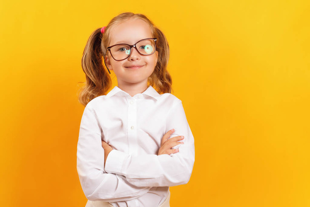 Cheerful little girl in glasses on a yellow color background. The child a schoolgirl in a white shirt folded her arms and looks at the camera. - Photo, Image