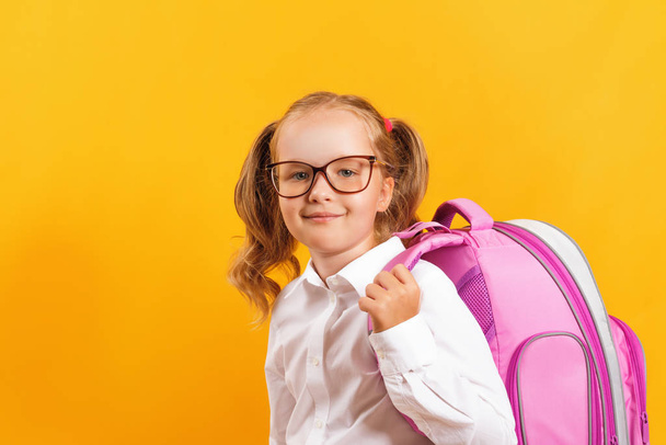 Little student girl in glasses on a yellow colored background. The child has a satchel behind his back. Education and school concept. - Photo, image
