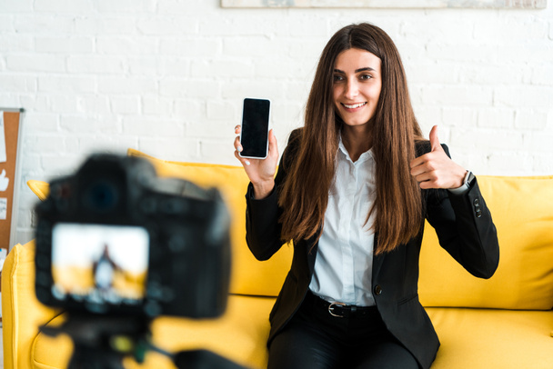 親指を表示し、デジタルカメラの近くに空白の画面でスマートフォンを保持する幸せな女性の選択的な焦点  - 写真・画像
