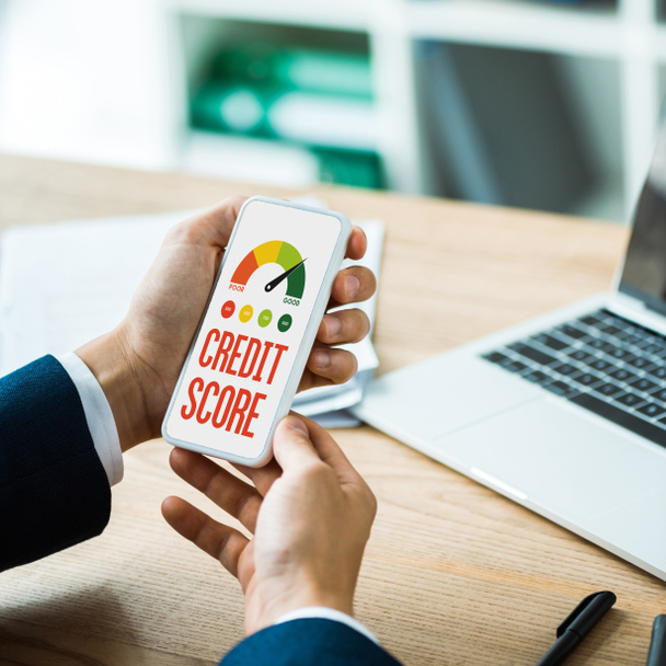cropped view of man holding smartphone with credit score lettering on screen near laptop and pen on table  - Фото, зображення