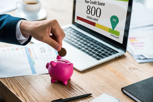 bijgesneden beeld van zakenman putting Coin in roze Piggy Bank in de buurt van laptop met uw credit score letters  - Foto, afbeelding