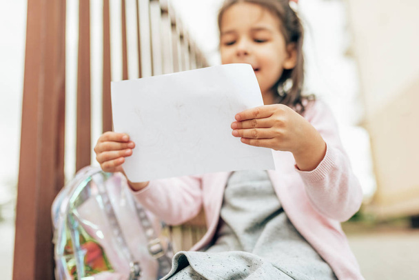 Image of cute little girl preschooler reading something on the paper sitting outdoors against blurred buildings. Happy kid pupil relaxing outside after preschool lessons. People and education concept - Photo, image