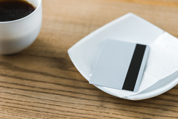πιστωτική κάρτα σε λευκή πλάκα με απόδειξη κοντά κούπα με καφέ σε ξύλινο τραπέζι  - Φωτογραφία, εικόνα