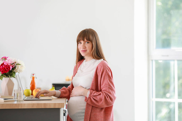 Beautiful pregnant woman with laptop in kitchen - Foto, afbeelding