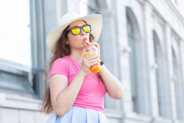Portrait of a cheerful positive young woman in a hat and glasses with juice in her hands walking around the city on a sunny warm summer day. The concept of carefree mood. - Photo, Image
