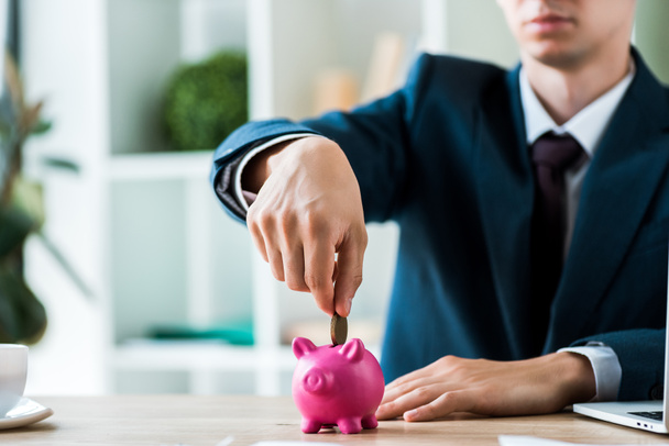 cropped view of businessman putting metallic coin into piggy bank near laptop - Photo, Image