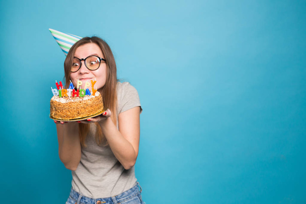Close up funny positive girl in glasses and greeting paper hat holding a happy birthday cake in her hands standing on a blue background. - Photo, Image