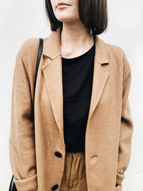 Herfst, herfst en mode concept. Een jonge vrouw met bruin haar, beige vacht, zwart overhemd, bruine broek staande tegen witte muur. Close-up foto met neutrale kleuren. - Foto, afbeelding