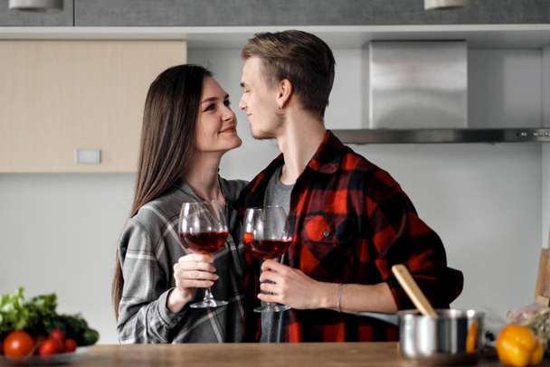 Beautiful young couple in plaid shirts in the kitchen preparing food and drinking red wine from glasses. - Φωτογραφία, εικόνα