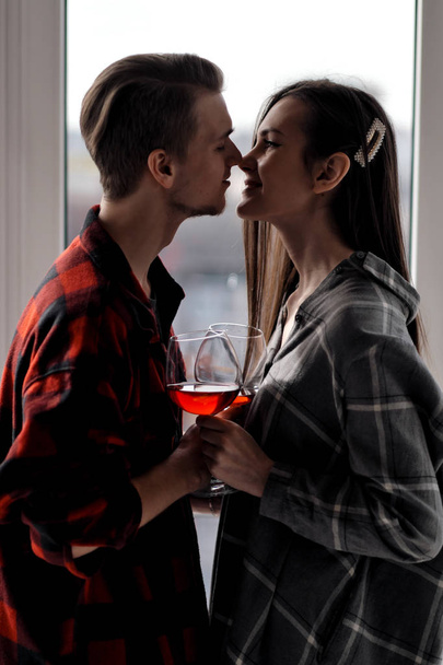 Young beautiful couple in plaid shirts on the balcony of a multistory building. Drink and taste wine, laugh and embrace. - Photo, Image