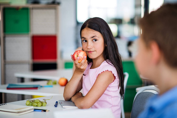 A small school girl sitting at the desk in classroom, eating apple. - Фото, зображення