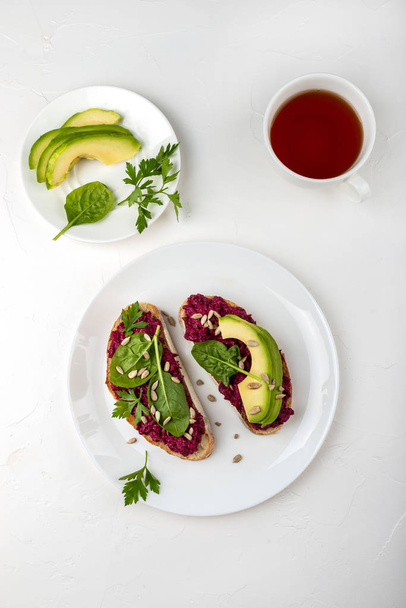 Ontbijt. Bruschettas met hummus van rode biet, avocado, spinazie en zaden op een witte plaat en kopje thee. Top View. - Foto, afbeelding