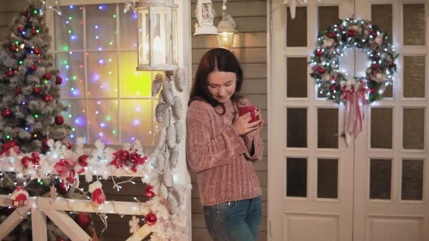 Young happy woman with red cup of coffee wearing knitted sweater standing at porch with christmas tree and christmass wreath at background. Brunette girl dreaming with copyspace at left - Filmagem, Vídeo