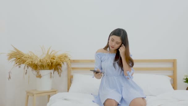 4K. beautiful Asian woman sitting on the bed listening to music with headphone from application on mobile phone and dancing with relax and happy emotion - Filmagem, Vídeo