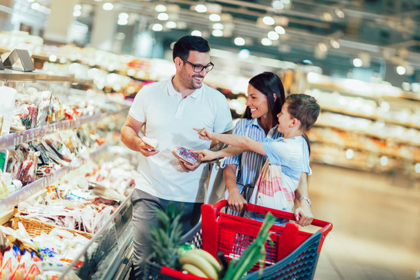 Onnellinen perhe lapsen ja ostoskorin kanssa ostamassa ruokaa ruokakaupasta
 - Valokuva, kuva