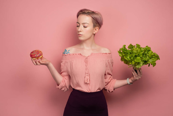 A girl with short pink hair, holding a salad and a donut in her hands, looks at him, on a pink background. - Foto, Bild