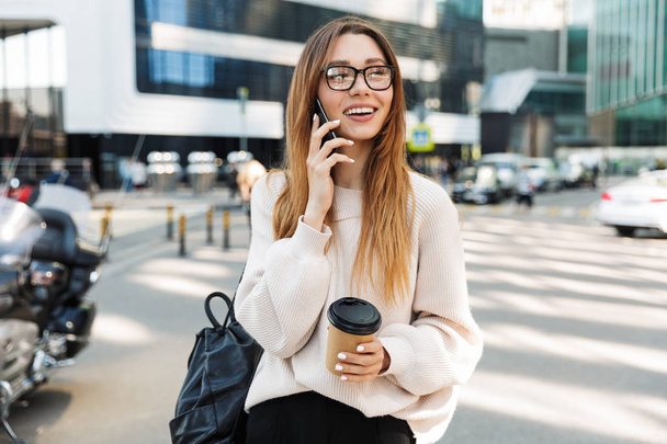 Photo of young caucasian woman talking on cellphone and holding paper cup while walking in big city street - Фото, зображення