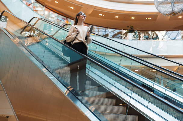 Full length photo of beautiful pretty woman smiling and holding clipboard while going down escalator in building indoors - Fotografie, Obrázek