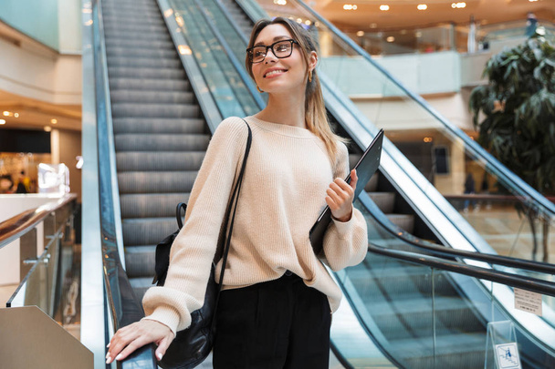 Photo of cute young woman smiling and holding clipboard while going down escalator in building indoors - Фото, зображення