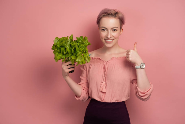 Ein Mädchen mit kurzen rosafarbenen Haaren lächelt fröhlich in die Kamera, hält einen Salat in der Hand und zeigt einen Daumen auf rosa Hintergrund. - Foto, Bild