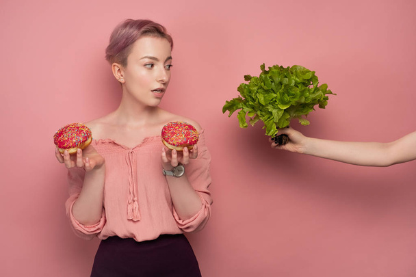 Uma menina com cabelo curto em uma blusa segura donuts em suas mãos, e olha para a salada na mão estendida, em um fundo rosa.
. - Foto, Imagem