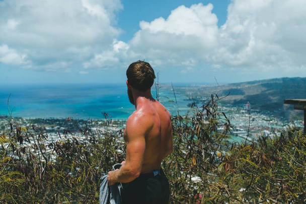 Closeup of a fit shirtless male standing on a high cliff with greenery and amazing clouds - Foto, Imagen