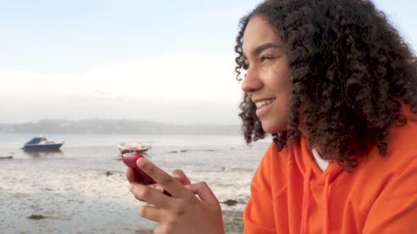 Beautiful mixed race African American girl teenager young woman wearing orange hoodie, sitting on a sea front using her smart cell phone for social media messaging or texting - Πλάνα, βίντεο