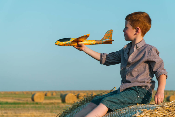 The red-haired boy plays with a toy airplane on a wheat field with bales - Fotografie, Obrázek