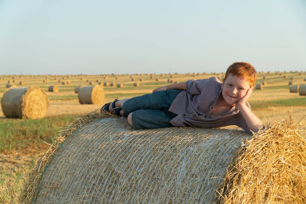 The red-haired boy lies on top of a straw bale on a wheat field - Фото, изображение