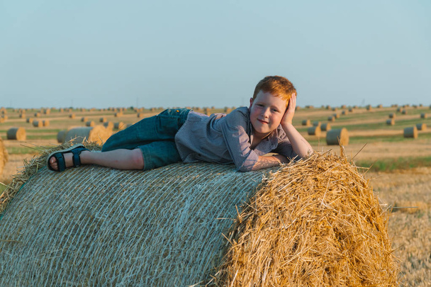The red-haired boy lies on top of a straw bale on a wheat field - Φωτογραφία, εικόνα