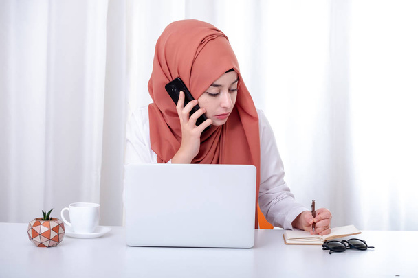 Musliminainen työskentelee tietokoneen, hijabin muslimitytön kanssa toimistossa ja hän puhuu matkapuhelimella, tuella, yhteyshenkilöllä tai asiakaspalvelukonseptin valokuvassa
 - Valokuva, kuva