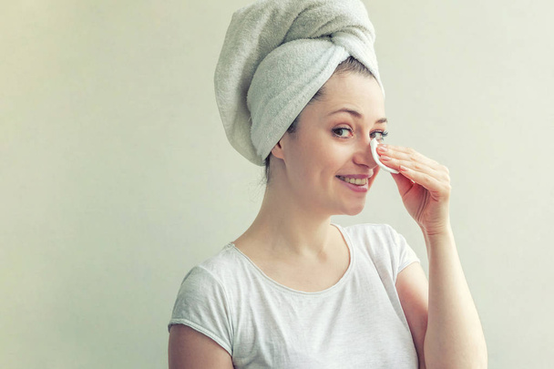 Усміхнена жінка в рушнику на голові з м'якою здоровою шкірою, що видаляє макіяж з ватною прокладкою ізольовано на білому тлі
 - Фото, зображення