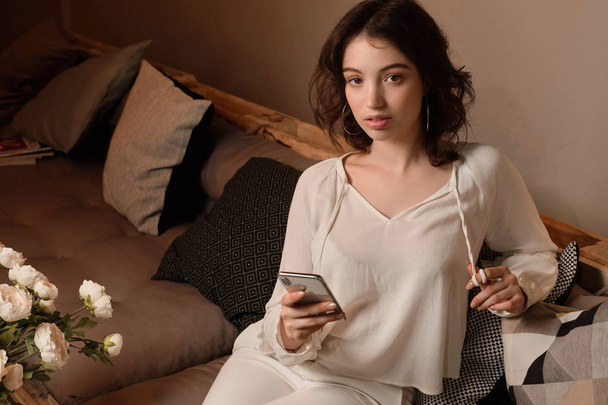 Retrato de una chica rizada de cabello oscuro en una blusa ligera sentada entre almohadas con un teléfono
. - Foto, Imagen