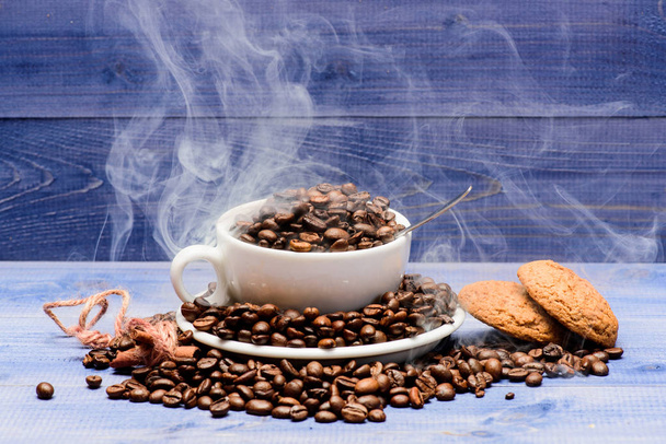 Coffee for inspiration and energy charge. Degree of roasting grain. Cafe drinks menu. Fresh roasted coffee beans. Cup full coffee brown roasted beans white clouds of smoke blue wooden background - Photo, Image