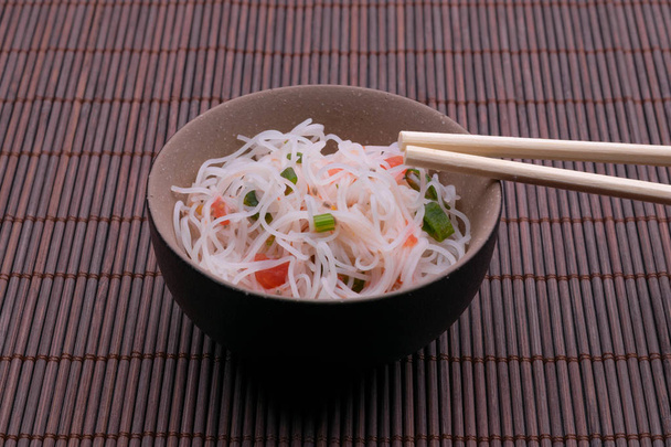 Ensalada asiática con fideos de arroz y verduras. Fideos de arroz cocidos estilo tailandés en un tazón negro y bronw con palillos aislados sobre fondo de madera
 - Foto, Imagen