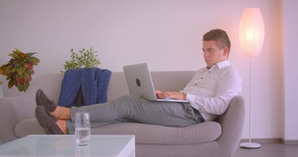 Closeup portrait of adult caucasian businessman using laptop sitting laidback on couch being sleepy and yawning in office indoors - Imágenes, Vídeo