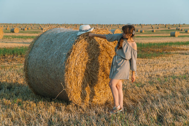 Hermosa joven con el pelo largo y sombrero posando en un campo de trigo cerca de fardos de heno
 - Foto, Imagen