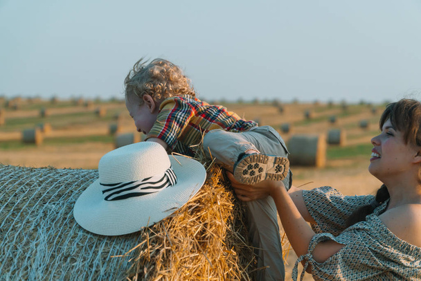 Young mother playing with her little son near a straw bale on a wheat field - Photo, image